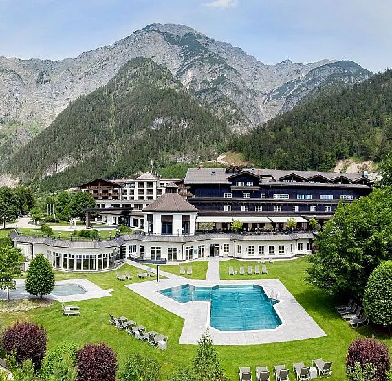 Luxury mountain resort with large pool, manicured gardens, and forested mountains in the background under a clear blue sky.