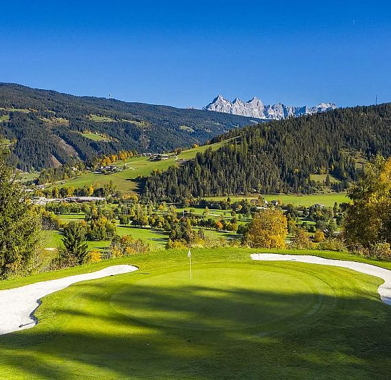 Golf course with green grass and sand bunkers, set against a mountain backdrop and blue sky.