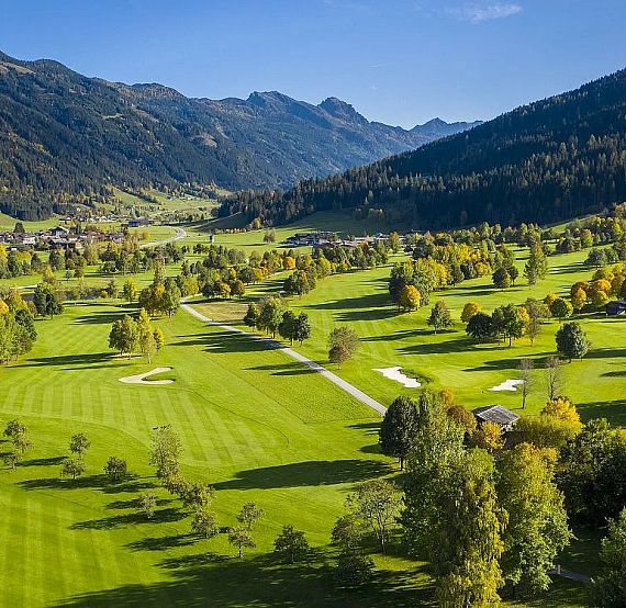 Aerial view of a golf course in a green valley with mountains, trees, fairways, and blue sky. Serene and picturesque landscape captured in the image.
