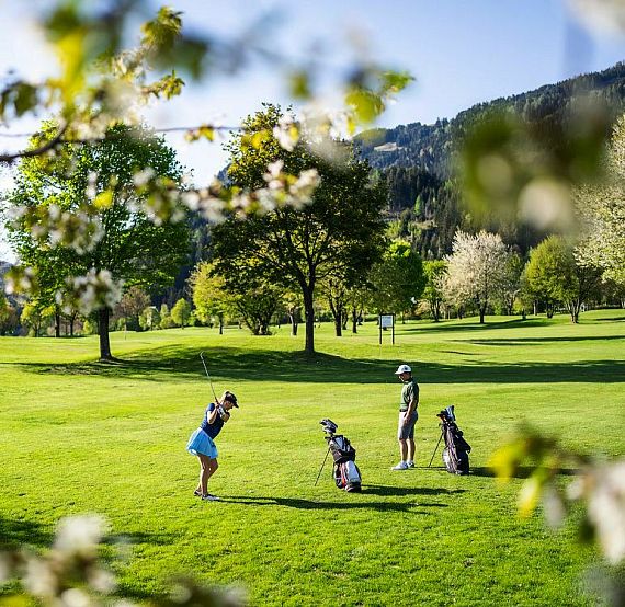 Drei Golfer auf einem grünen Golfplatz, umgeben von Bäumen und blühenden Zweigen, in einer sonnigen und landschaftlich reizvollen Umgebung.