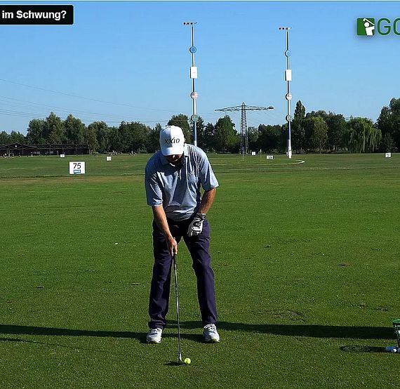 A person at a driving range preparing to swing at a golf ball, surrounded by green grass and golf equipment under a clear blue sky.