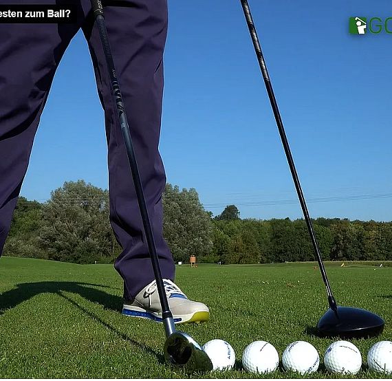 A golfer stands on a green field with several golf balls and a club, showing proper stance for a swing. Trees and blue sky are in the background.
