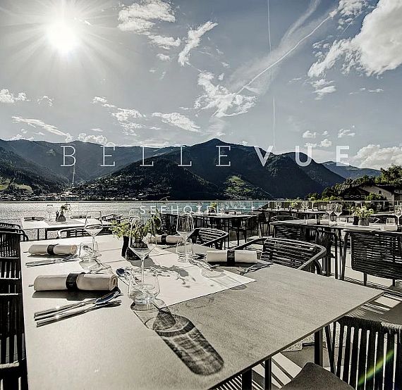 Outdoor restaurant by a lake with mountains, under a sunny sky, featuring tables and chairs set for dining.