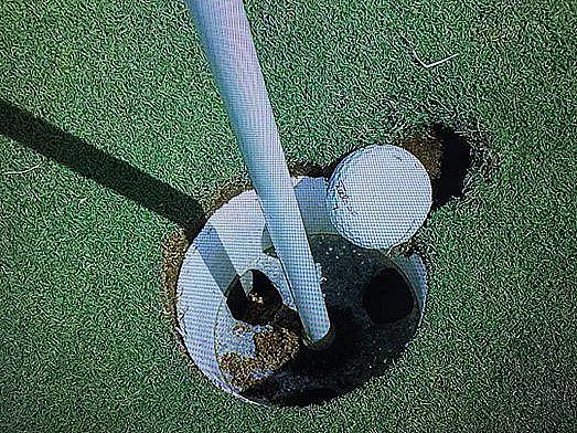 A golf ball balances on the edge of a golf hole on a green, creating a tense moment. The close proximity hints at a near miss or a successful putt.