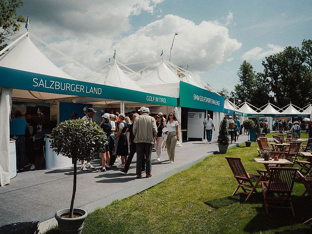 Besucher schlendern über ein Freiluft-Event mit weißen Zeltdächern und blaugrünen Schildern. Im Vordergrund stehen kleine Bäume und Holzmöbel auf dem Rasen.