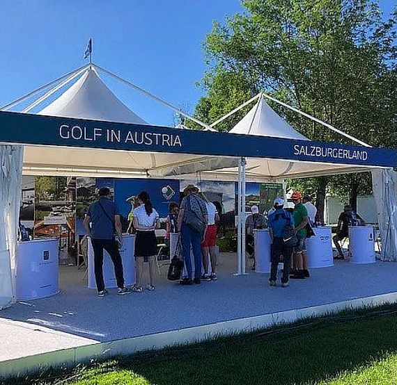 People gather under a white tent labeled 'Golf in Austria' and 'Salzburgerland,' set outdoors in a sunny park with green grass and trees.