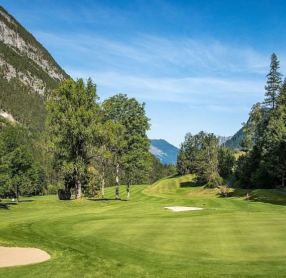 A golf course with sand bunkers, manicured greens, surrounded by mountains and trees under a clear blue sky.