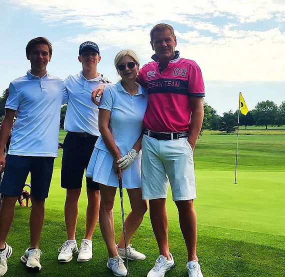 Four people posing on a golf course near the green. They're dressed in sporty attire, holding golf equipment, with a flag in the background. It's a sunny day.