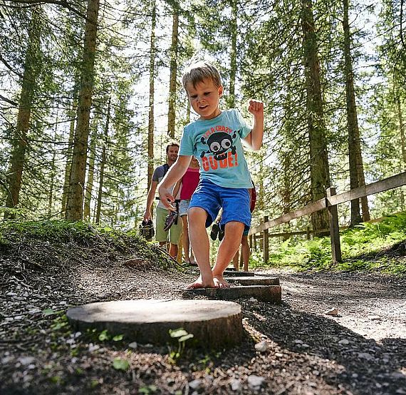 A boy wearing a blue shirt and shorts jumps on wooden discs on a forest path. Several adults follow him. Tall trees surround them, creating a sunny woodland scene.