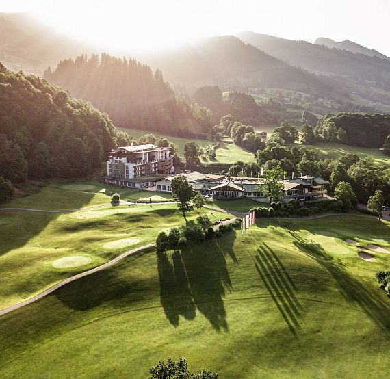 Aerial view of an alpine resort with lush greenery, surrounded by mountains under sunlight casting long shadows, creating a picturesque and serene atmosphere.