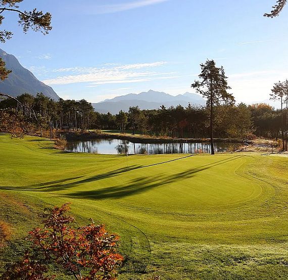 A picturesque golf course with green fairways, trees, and a pond, set against a backdrop of mountains and a blue sky.