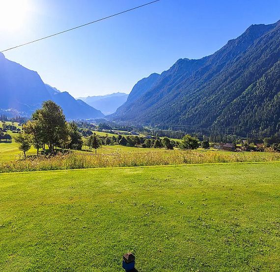 A wide green valley with towering mountains under a bright blue sky. The landscape is tranquil, featuring lush lawns and a backdrop of forested peaks.
