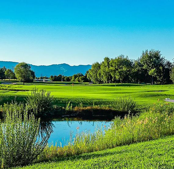 Lush green golf course with a pond and trees under a clear blue sky.