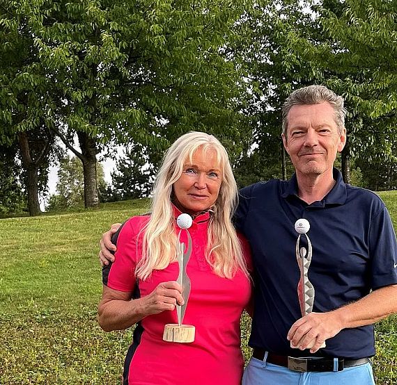 A man and a woman standing outside holding trophies in front of a background with trees and grass. The woman wears a pink shirt, and the man a dark polo.