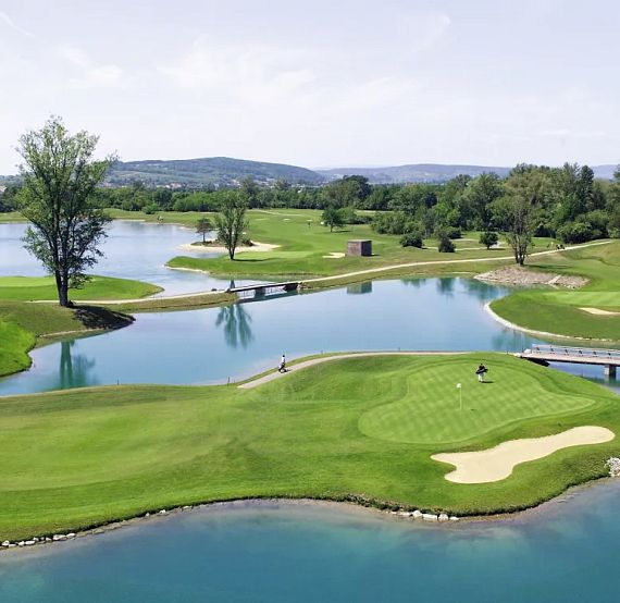 Aerial view of a golf course with clear water lakes, green fairways, sand bunkers, and trees, set against a backdrop of distant hills under a blue sky.