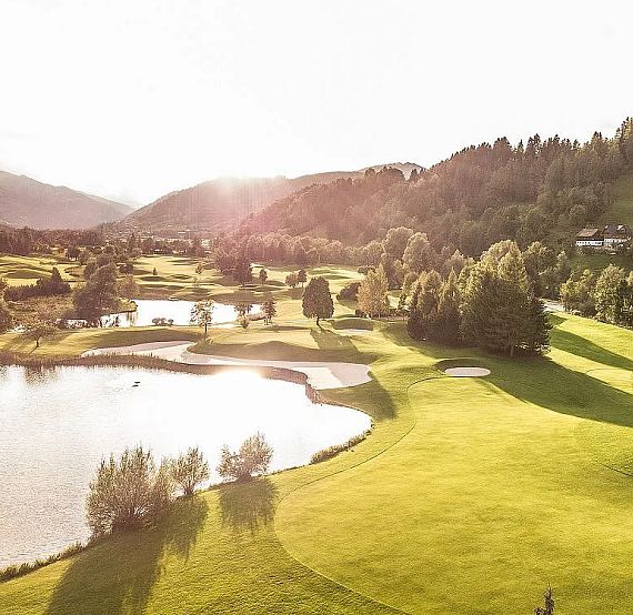Aerial view of a green golf course with a lake, trees, and hills under a setting sun, creating a serene and picturesque landscape.