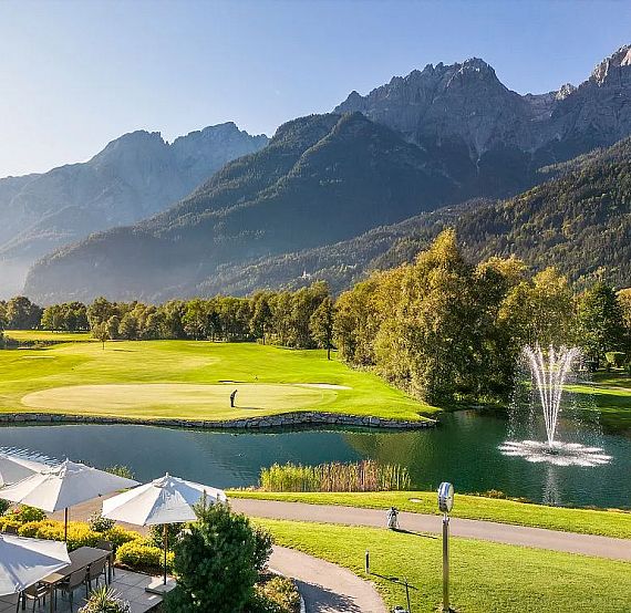 A scenic golf course with mountains in the background, a water feature with a fountain, and a golfer on the green, set amidst lush trees and landscape.