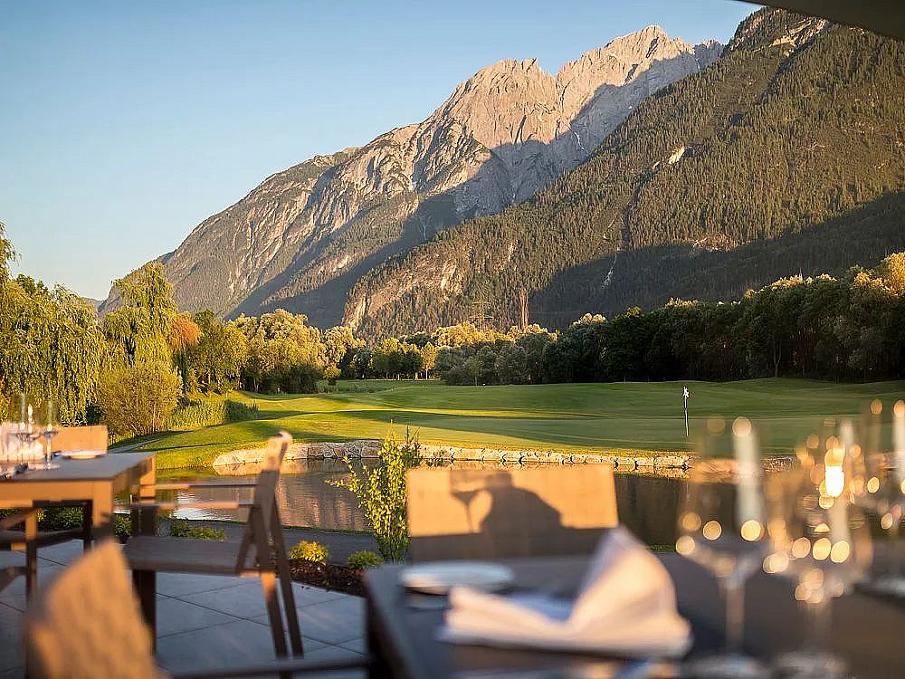 Ein Restaurant mit Blick auf einen Golfplatz, umgeben von majestätischen Bergen bei Sonnenuntergang. Die Tische im Vordergrund sind elegant gedeckt.