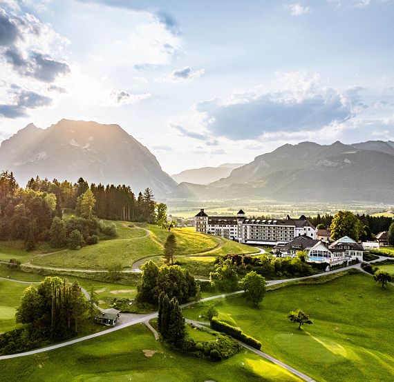 A scenic view of a verdant valley with a modern building, lush trees, and mountains under a partly cloudy sky with sunlight streaming through.