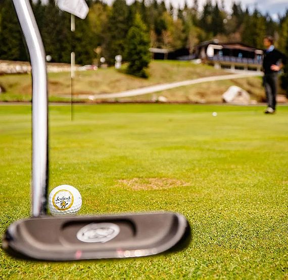 Close-up of a golf putter and ball on a green. A golfer stands in the background ready to take a putt. The background is lush with trees and hills.