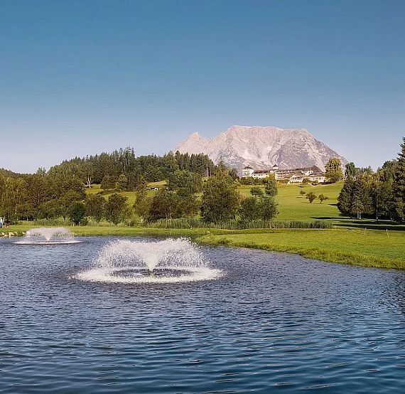 A lake with two fountains, surrounded by greenery and trees, with a mountain in the background under a clear sky.