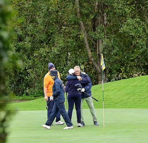 Golfers on a course celebrate as two hug near a flag. Trees and greenery surround the scene, creating a lively outdoor backdrop.