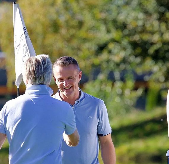 Two men in polo shirts outdoors; one smiles, the other offers a handshake. Sunny background with greenery and a hint of a building.