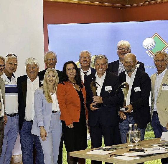 Group of people at a golf awards ceremony, some holding trophies, in a formal setting with a large screen in the background.