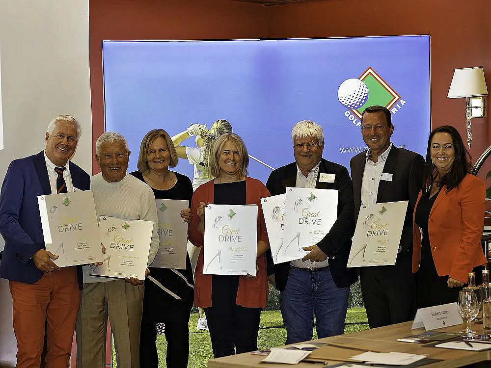 A group of people holding certificates stand smiling in front of a backdrop with a golf theme, suggesting a recognition event.