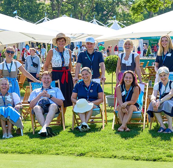 Group of people sitting on deck chairs under white umbrellas, smiling at a golf event, some in traditional costumes, with a green lawn and trees in the background.