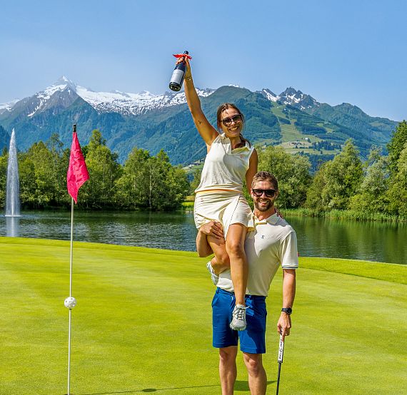 Ein Paar steht auf einem Golfplatz vor einer malerischen Bergkulisse. Die Frau sitzt auf den Schultern des Mannes und hält eine Flasche Champagner hoch.