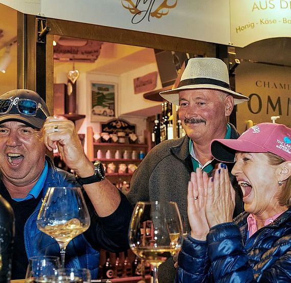 Three people in casual attire enjoying a moment of celebration in a wine shop, with glasses and bottles around them. They are smiling and cheering enthusiastically.