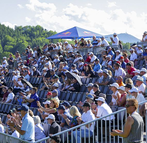 A crowded grandstand with people sitting, clapping, under a clear sky. Green trees and mountains are in the background. Some spectators wear hats.