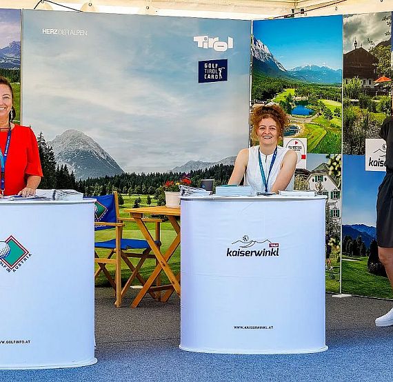 Three women stand at booths in a trade fair setting, each representing a different tourist region. The background shows scenic images of mountains and towns.
