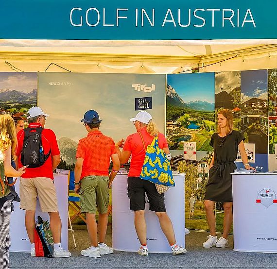 People standing at a booth under a banner that reads 'Golf in Austria', with promotional displays and a staff member present.