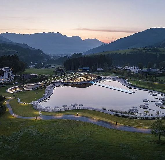 Modernes Hotel-Resort in alpine Landschaft eingebettet, umgeben von Hügeln und Bergen. Eine zentrale Wasserfläche spiegelt den farbenfrohen Abendhimmel wider.