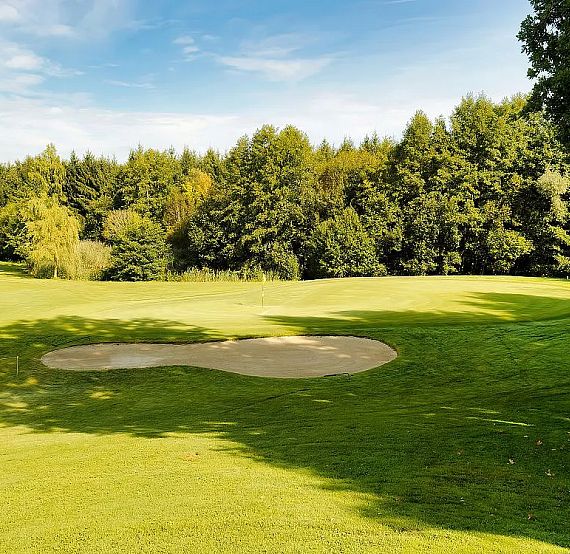A golf course with a sand trap on lush green grass, surrounded by trees and a clear blue sky.