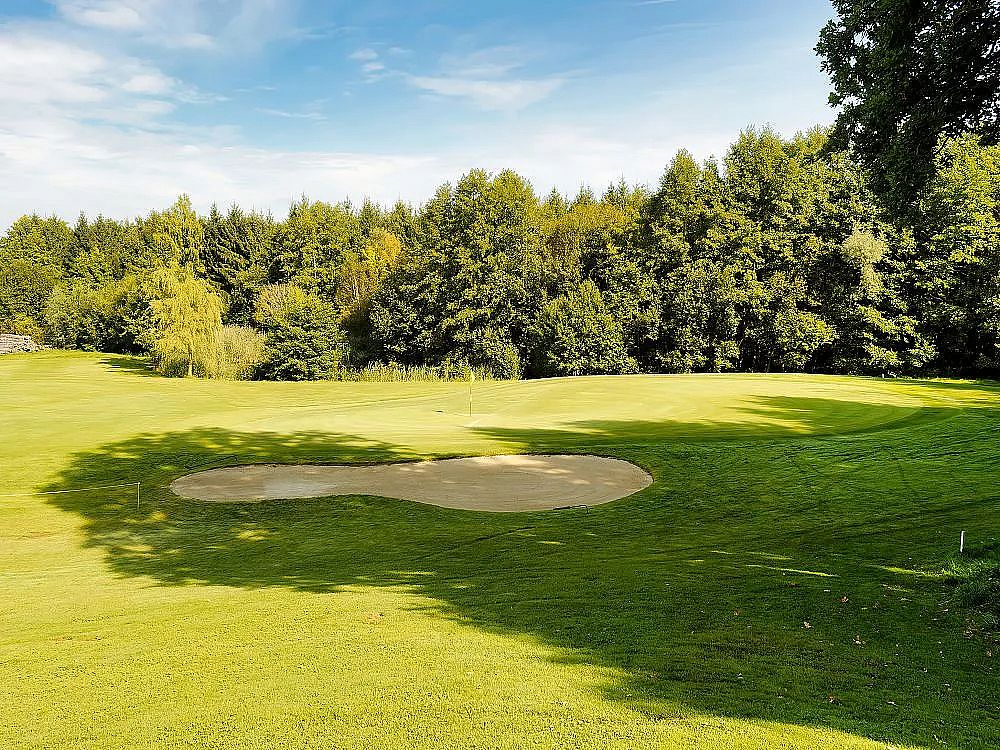 A golf course with a sand trap on lush green grass, surrounded by trees and a clear blue sky.