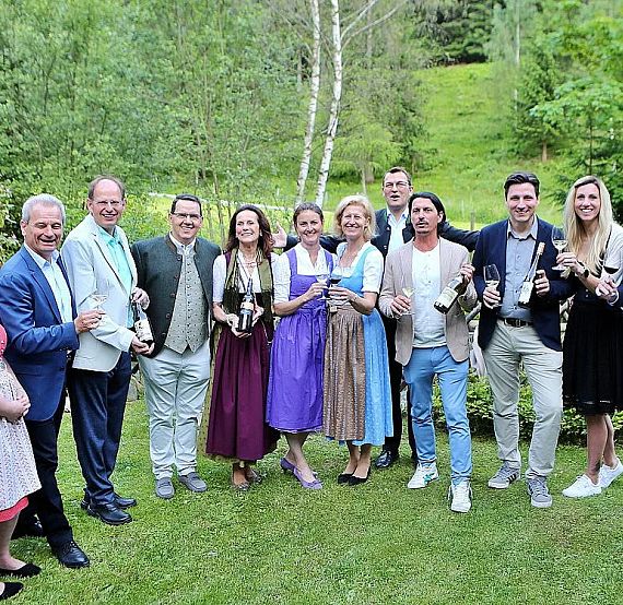 A group of people stands outdoors in lush, green surroundings, each holding a beverage bottle, dressed in formal and traditional attire, suggesting a celebration.