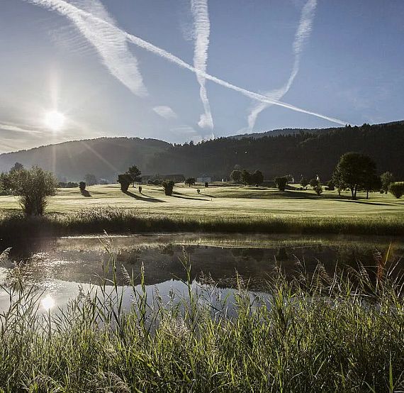 Sun rises over a tranquil golf course with green grass, scattered trees, and a pond reflecting the sky. Contrails create patterns against the clear blue sky.