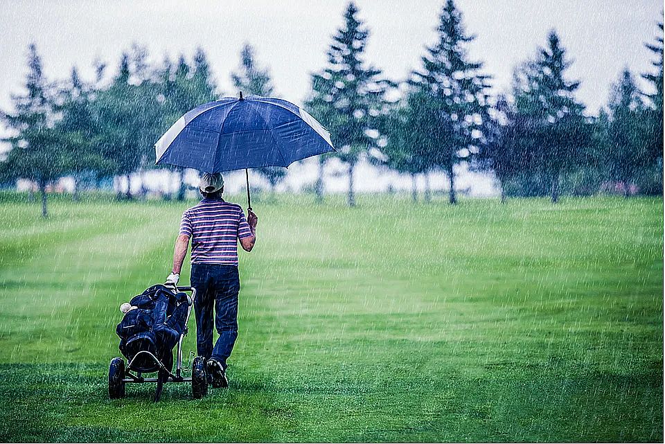 Person on a golf course holding an umbrella, walking in the rain with a golf cart, surrounded by lush greenery and distant trees.