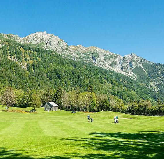 A lush, sunlit golf course with green fairways set against a backdrop of forested mountains, under a clear blue sky. A small building and trees are visible.