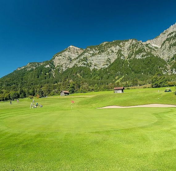 Lush green golf course with sand bunker, surrounded by mountains and a clear blue sky.