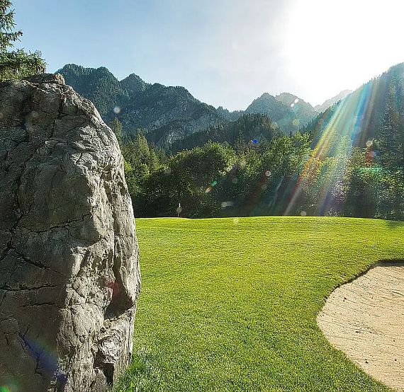 Large rock on green grass with sand trap and mountains in background under bright, sunny sky.
