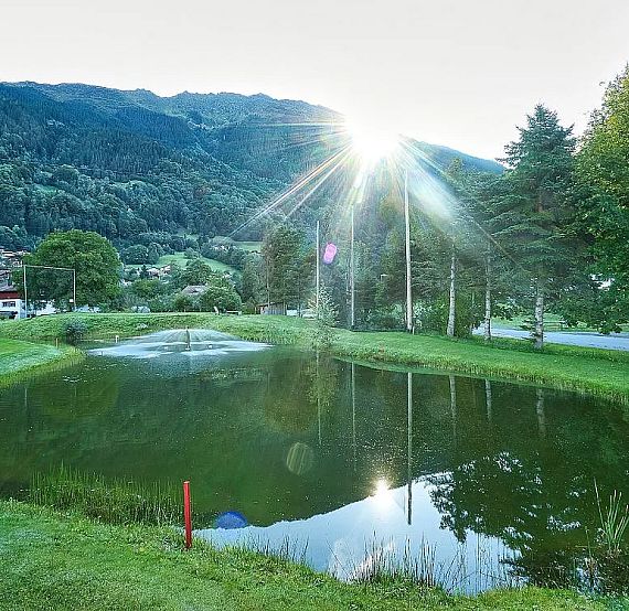 A tranquil pond with reflections of trees, set against a mountain backdrop. Sun rays filter through trees, adding a serene glow to the peaceful landscape.