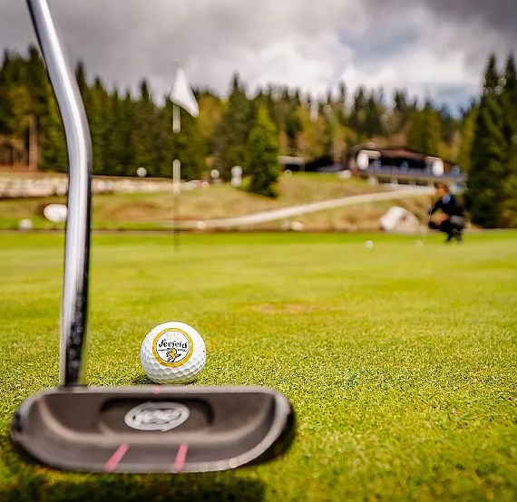 Close-up of a golf ball on a green, with a putter nearby. Trees and a clubhouse are in the background under a cloudy sky.