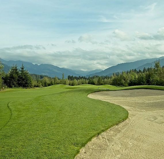Lush golf course with green grass, a sand bunker, and surrounding mountains under a cloudy sky, creating a serene and picturesque outdoor scene.