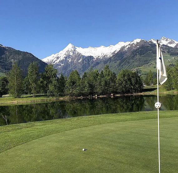 A serene golf scene with a green, a flag, a lake, trees, and snow-covered mountains under a blue sky.