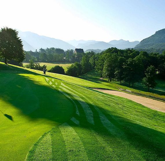 Ein grüner Golfplatz bei Sonnenaufgang, umgeben von Bäumen und Hügeln im Hintergrund. Die Landschaft wirkt ruhig und friedlich mit strahlend blauem Himmel.