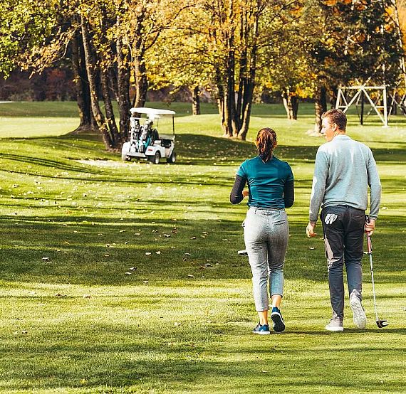 Two people walking on a golf course in autumn, with trees losing leaves and a golf cart in the background. The scene is peaceful and green.
